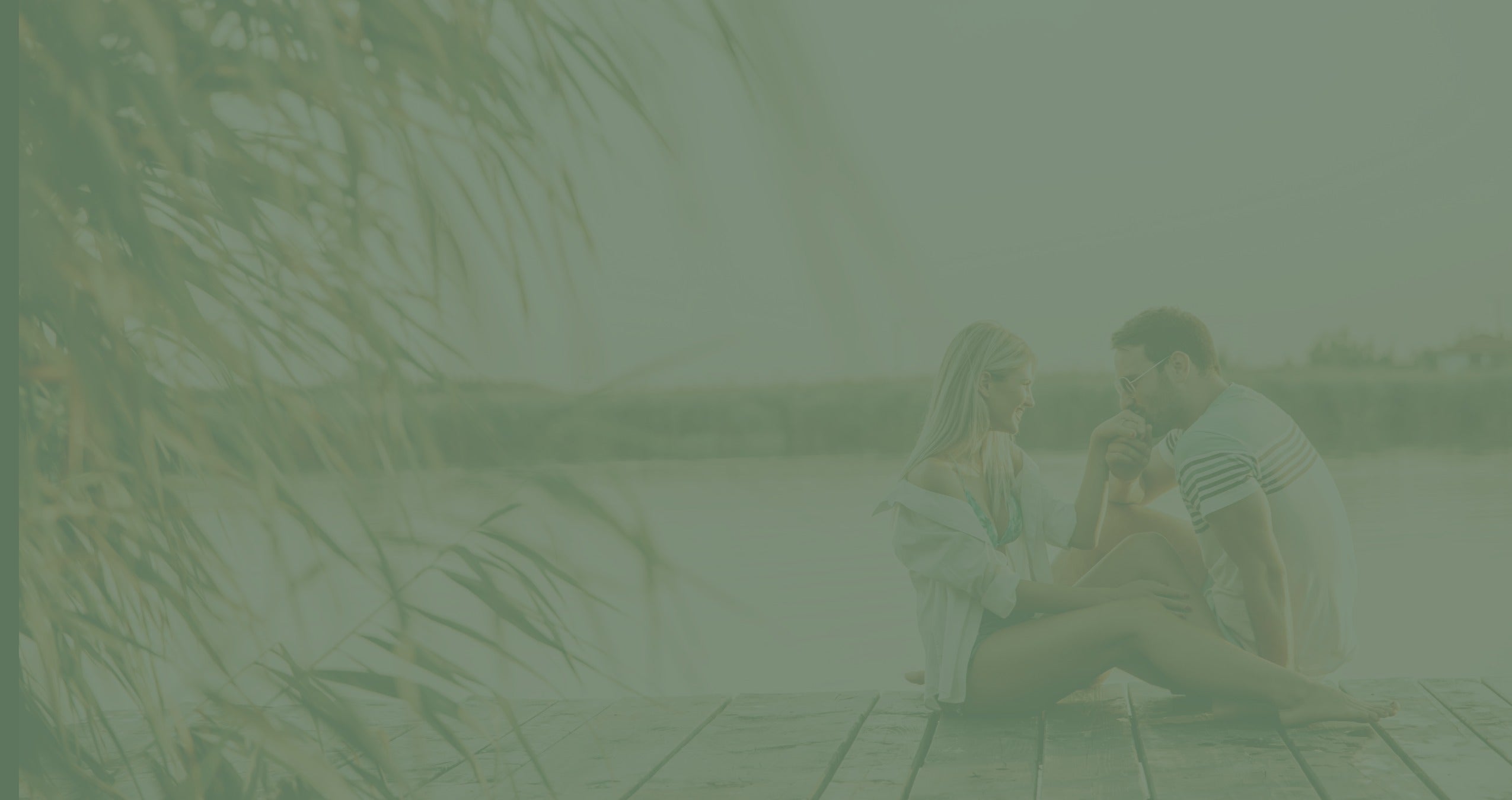 A muted green-toned lifestyle photograph of a couple sitting together on a wooden dock by the water, with reeds in the foreground.