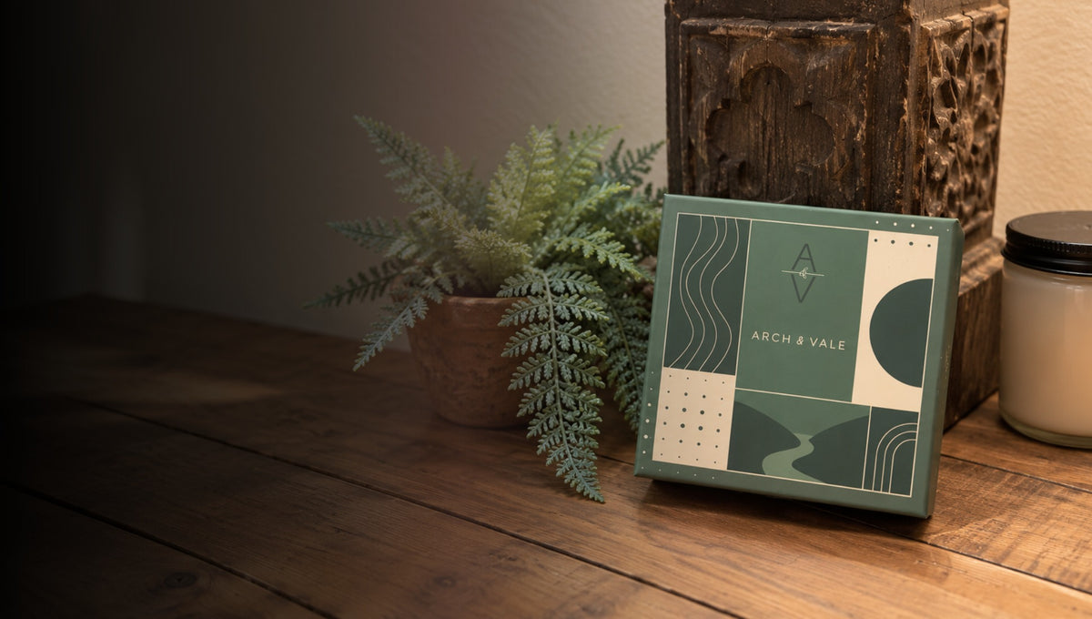 A square decorative product box for Arch & Vale sits on a wooden table beside a potted fern, with a rustic wooden object and a candle in the background.