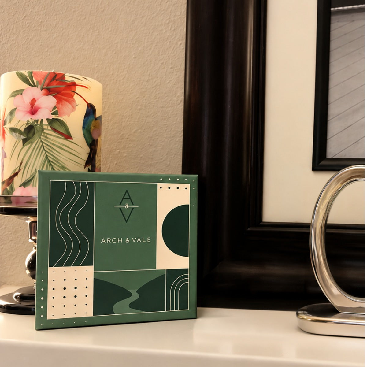 A square decorative box or package labeled ARCH & VALE sits on a tabletop in a styled interior setting. The front of the package has an abstract geometric pattern in dark green and cream, with a small mountain-and-tree emblem above the brand name.