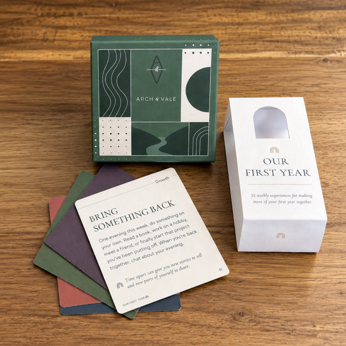 Arch & Vale 'Our First Year' card deck set on a wooden tabletop, featuring a green geometric patterned box, a white card deck packaging sleeve, and several colorful cards fanned out.